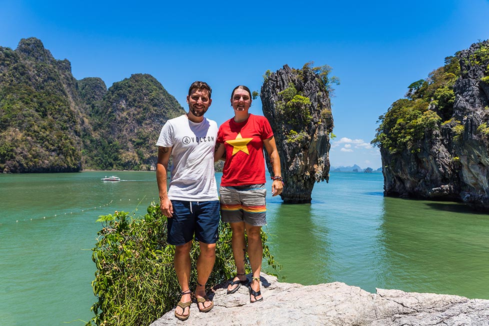 Nic and shorty standing on a rock with James Bond Island, near Phuket in Thailand behind them. It is a sunny day with a blue sky and green-blue sea water Thailand backpacking packing list guide.