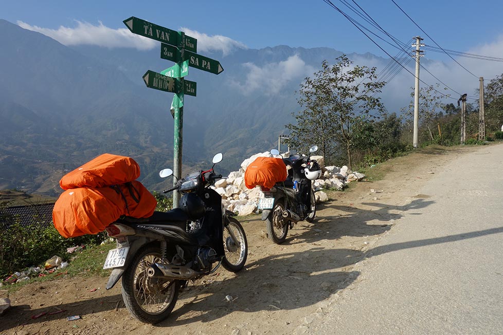 Two motorbikes with bags attached are parked up next to a sign for Sapa and Ta Van with mountains and rice paddies below in Vietnam