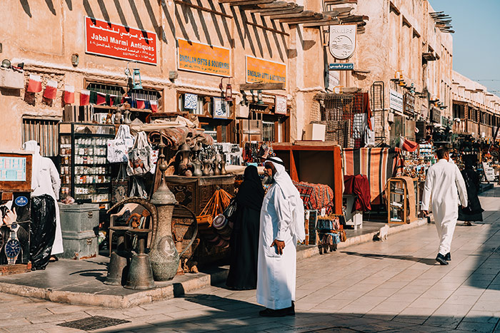 Qatari men walking along the streets of the souk outside an antique shop selling traditional copper pots and jugs on a sunny day in the souk in Doha, Qatar