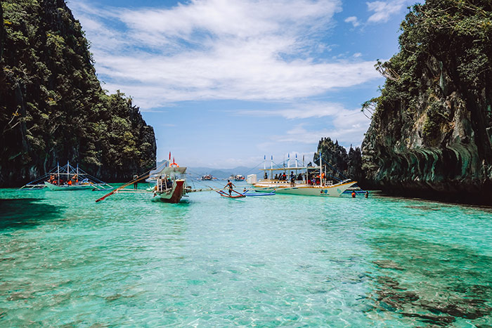 clear green - blue water in a narrow opening with high limestone cliffs on either side with traditional boats sailed down the middle near El Nido, Philippines