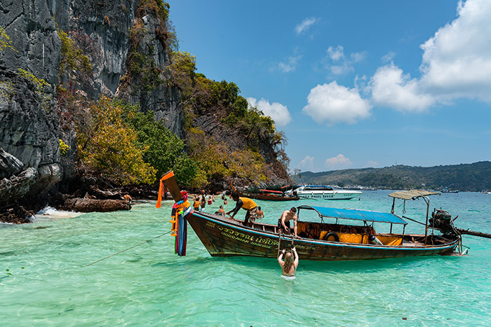 A traditional long tail boat pulled up at a beach next to a limestone cliff near Phuket Phi Phi Thailand with beautiful clear blue green sea around it