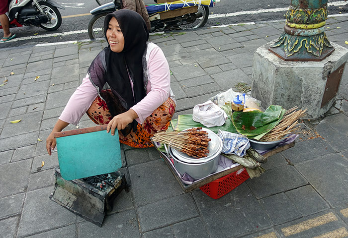 A muslim woman sitting on the street in Yogyakarta, Indonesia selling street food she is cooking over a small grill