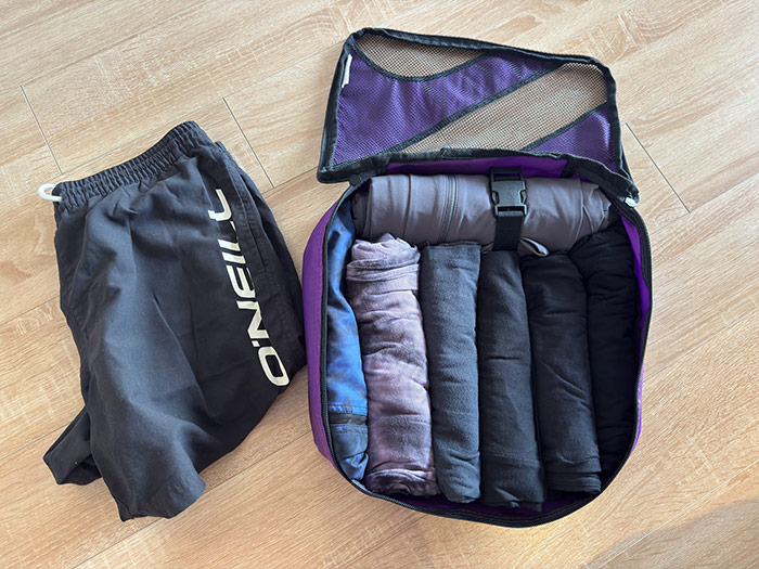 Nic's clothing packing cube which is open and showing 1 pair of shorts, 5 t-shirts, and 1 pair of trousers inside. With another pair of shorts next to it.