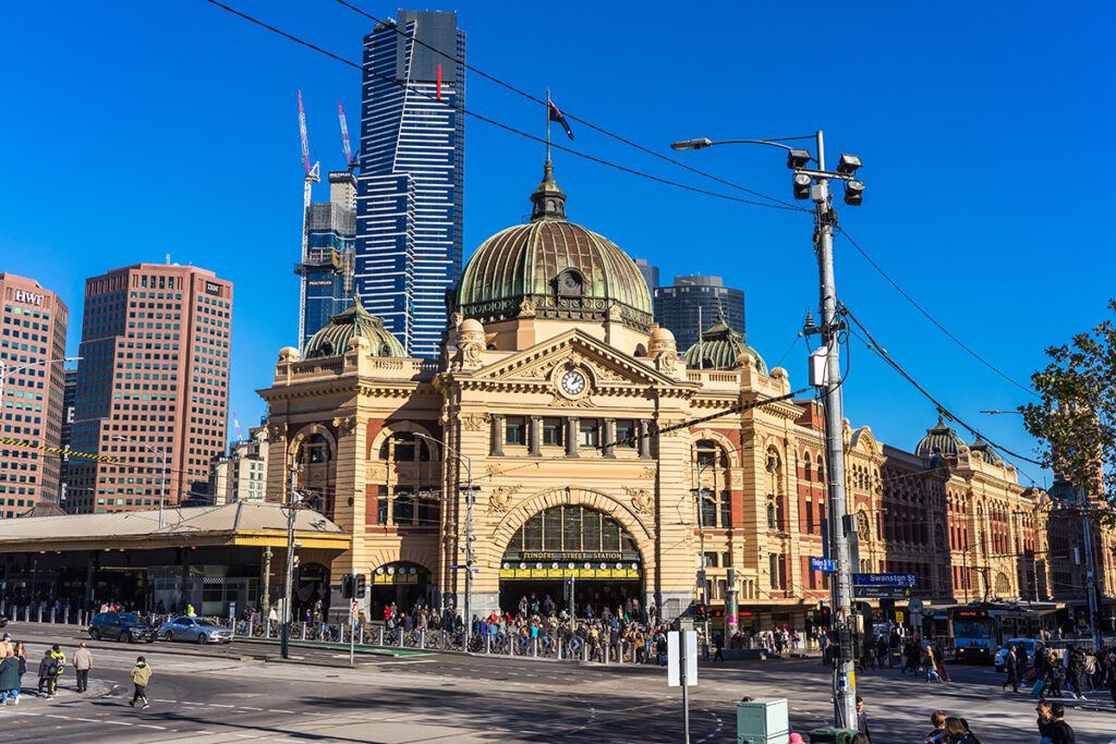 The iconic Flinders Street train station with pedestrians crossing the road in the foreground on a sunny day. It's one of the best free things to do in Melbourne. 