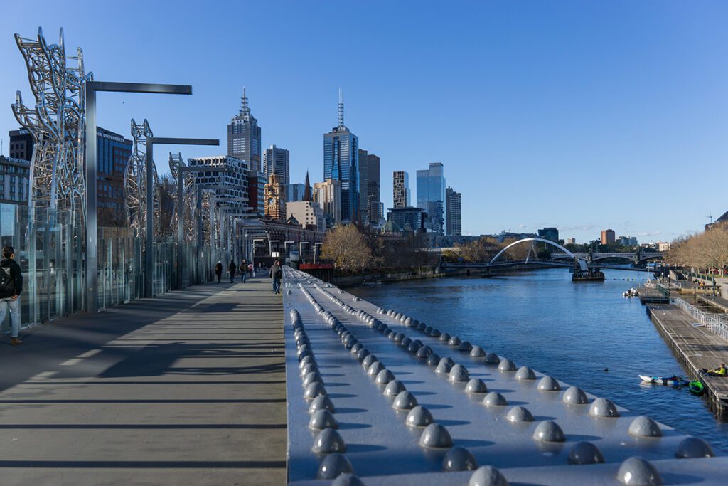 A walkway next to the Yarra River with tall buildings and bridges in the background.