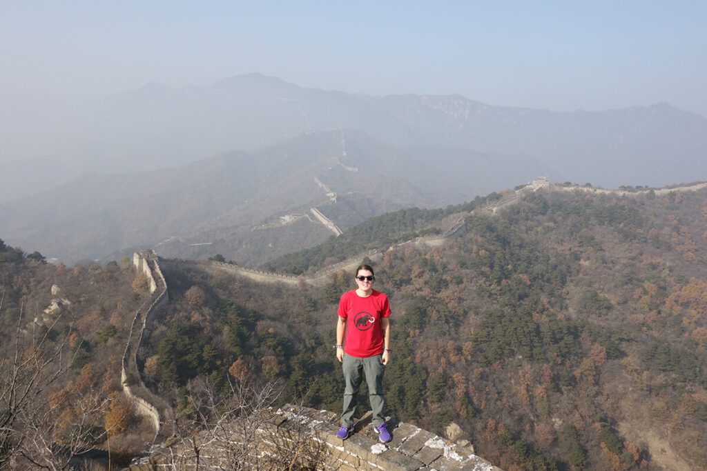 Nic standing on an undeveloped section of the Great Wall of China which stretches out behind them into the mountains that are covered in smog.