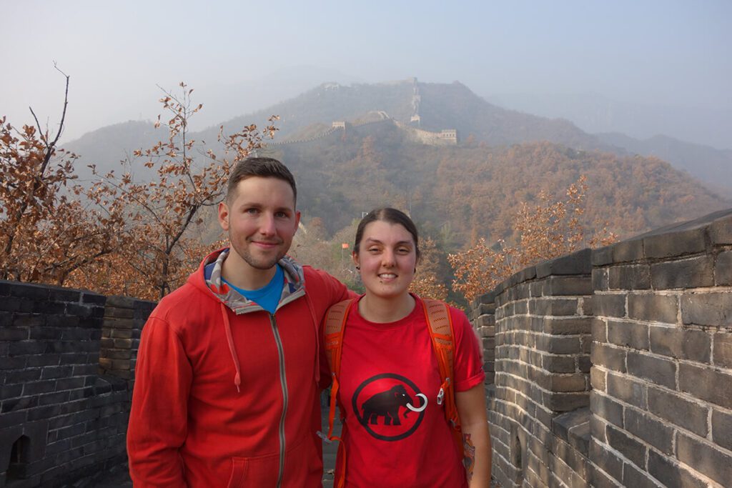 A picture of Nic and Shorty on the Great Wall of China with the wall behind them on a smoggy days, you can see mountains and autumn leaves