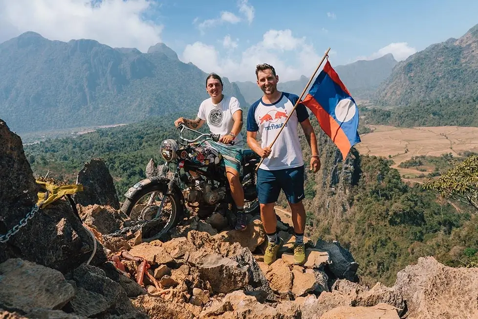 Nic and Shorty of Stories, Visions, Hope, Self standing on a summit in Laos and smiling broadly while holding a Laotian flag.
